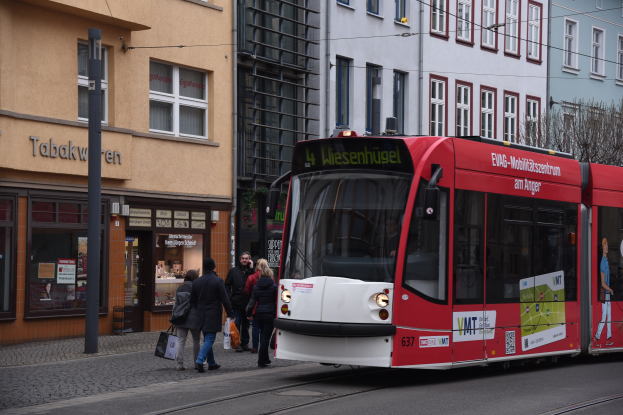 Eine rote und weiße Straßenbahn auf einer Stadtstraße mit Menschen, die gehen und Taschen tragen, Gebäude, Bäume und eine Stange im Hintergrund.