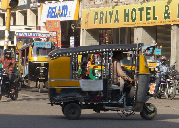 Eine belebte Straße mit verschiedenen Fahrzeugen, einschließlich eines Auto-Rikschas, und Menschen mit Helmen, mit einer Ampel im Vordergrund und Gebäuden mit Namensschildern und Rollläden im Hintergrund.