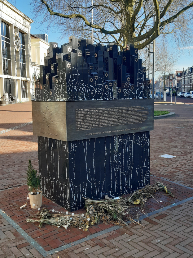 Ein Denkmal f√ľr Holocaust-Opfer steht auf einem Platz, mit einem Baum davor, einem Topf mit einer Pflanze, verstreuten trockenen Bl√§ttern, Geb√§uden, Fahrzeugen, Masten und B√§umen im Hintergrund.