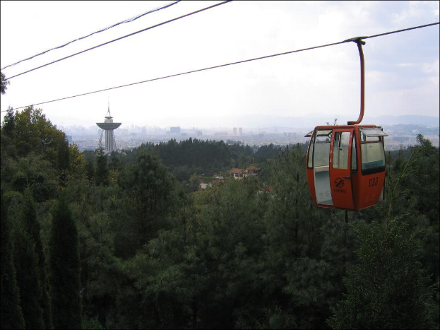 Eine Seilbahn fährt im Vordergrund mit Bäumen, Gebäuden, einem Turm und einer Stadtlandschaft im Hintergrund unter einem bewölkten Himmel.