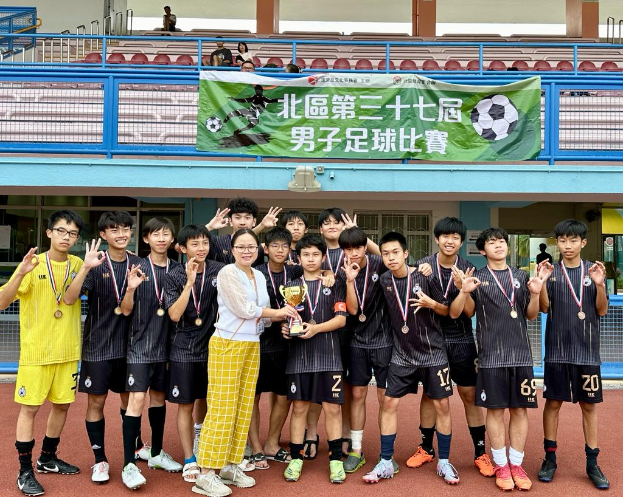 Eine Gruppe junger Männer in Fußballtrikots steht zusammen auf einem Feld, hält einen Pokal und trägt Medaillen, mit einem Gebäude im Hintergrund, das ein "Yokohama U-16 Jungenfußballteam"-Schild zeigt.