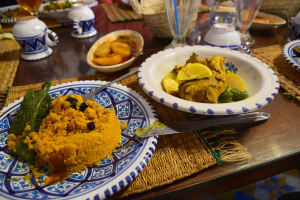 Ein Tisch mit Tellern, Wein- und Schüsseln, Schalen, Besteck und Matten, mit Menschen im Hintergrund bei einem traditionellen marokkanischen Essen.