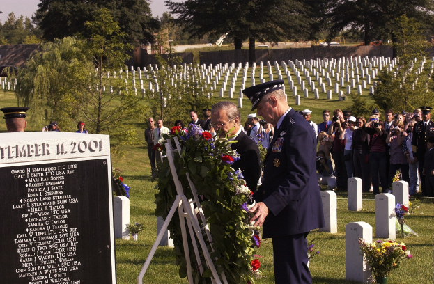 Eine Gruppe von Menschen versammelt sich um ein Grab auf einem Friedhof, wo ein Mann in einer Militäruniform einen Kranz niederlegt; Blumenbouquets, ein Textschild und umliegende Vegetation sind sichtbar, mit einem Haus, Bäumen und einem bewölkten Himmel im Hintergrund.