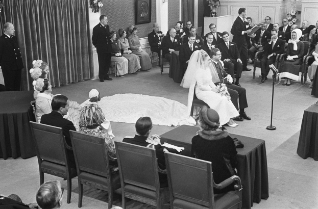 Schwarzes und weißes Foto einer Hochzeitszeremonie mit Menschen auf Stühlen und Stehplätzen, ein Mann mit Mikrofon, eine Frau mit Blumenstrauß, ein Podium mit Mikrofon auf der rechten Seite, Blumengestecke und ein Vorhang auf der linken Seite, ein Foto Rahmen an der Wand und eine Tür im Hintergrund.