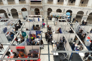 Eine große Gruppe von Menschen versammelt sich in einem Raum, der mit Ständen, Bannern, Tischen, Stühlen und Luftballons gefüllt ist, mit einem Gebäude im Hintergrund, das Bögen, Säulen, Geländer und einen Bildschirm aufweist.