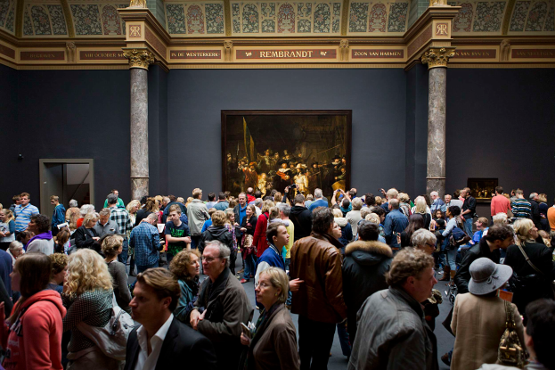 Eine große Gruppe von Menschen in einem Raum mit einem Foto auf der Wand, Säulen, einer Tür und einer Decke mit Lampen, einige halten Taschen, im Stedelijk Museum in Amsterdam.