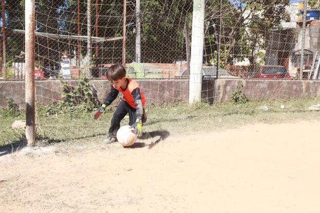 Junge in Fußbekleidung spielt Füssball auf einem Schotterfeld mit Gras, Pflanzen, Pfosten, einem Zaun, einer Wand, Bäumen, Fahrzeugen, Gebäuden und einer bewölktem Himmel im Hintergrund.