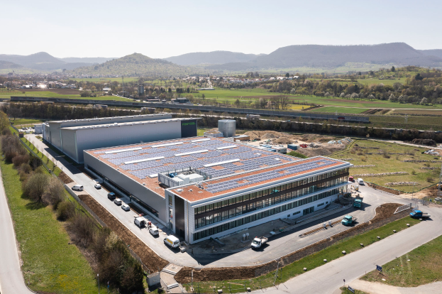 Eine Luftaufnahme eines großen Fabrikgebäudes mit Solarpaneelen auf dem Dach, umgeben von Fahrzeugen, Bäumen, anderen Gegenständen und Hügeln mit einem klaren blauen Himmel im Hintergrund, die erste Fabrik in Deutschland, die mit Solarenergie gebaut wurde.