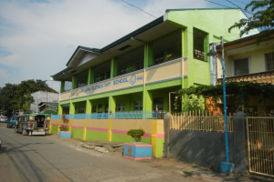 Schulgebäude mit grüner und gelber Fassade, Fahrzeugen auf der Straße, Topfpflanzen, einem Zaun, einem Tor, einem Straßenpfahl, Bäumen, Stromleitungen und einem bewölkten Himmel.