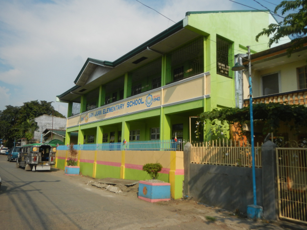 Schulgebäude mit grüner und gelber Fassade, Fahrzeugen auf der Straße, Topfpflanzen, einem Zaun, einem Tor, einem Straßenpfahl, Bäumen, Stromleitungen und einem bewölkten Himmel.