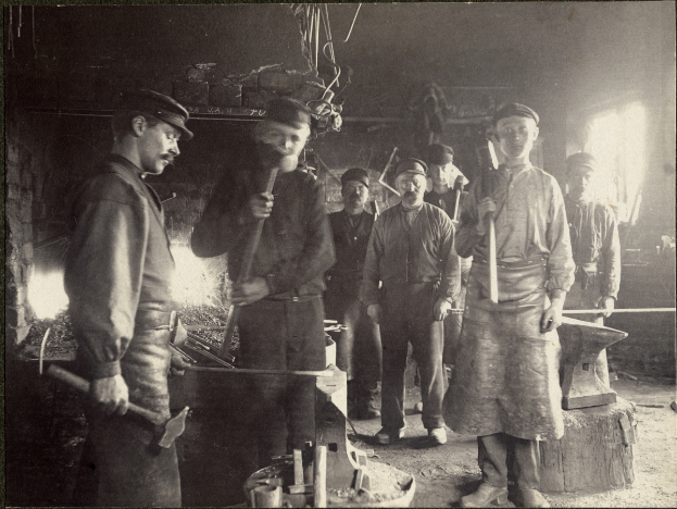 Eine Gruppe von Männern in einer Schmiede, jeder hält einen Hammer in der Hand, auf einem Schwarz-Weiß-Bild.