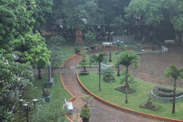 Ein grüner Park mit Bäumen, Pflanzen, Gras, Laternen, Bänken und Skulpturen sowie einem Gebäude im Hintergrund.