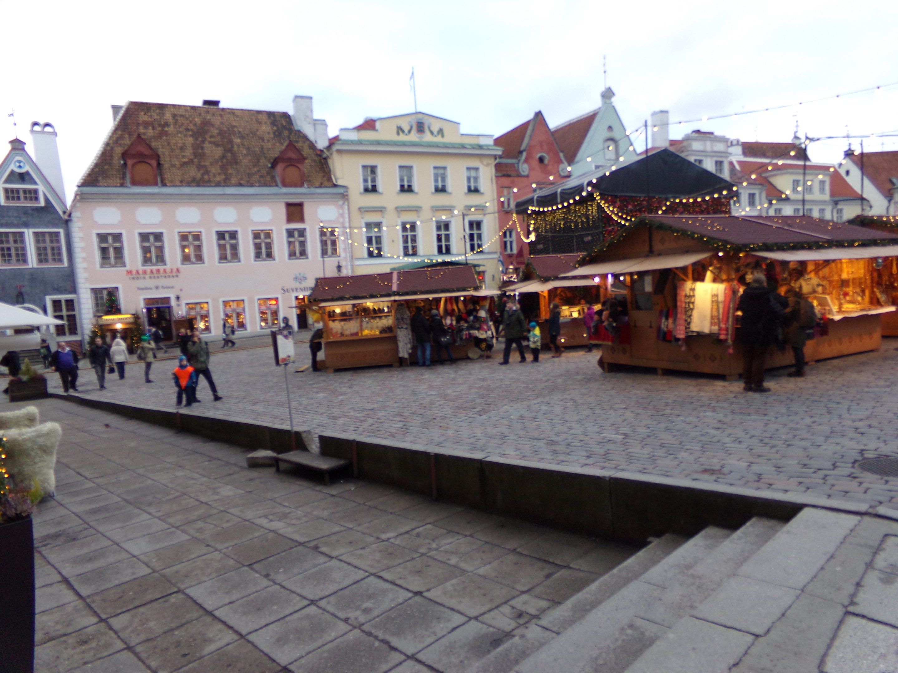 Ein geschäftiger Weihnachtsmarkt in Tallinn, Estland, mit Menschen um geschmückte Stände, festlicher Beleuchtung, bewölktem Himmel, Treppen, die hinaufführen, und Topfpflanzen im Bereich.