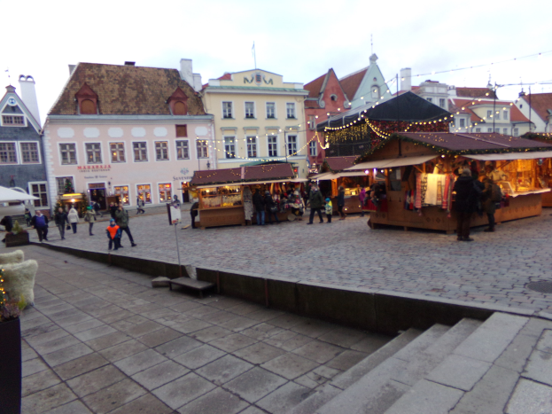 Ein geschäftiger Weihnachtsmarkt in Tallinn, Estland, mit Menschen um geschmückte Stände, festlicher Beleuchtung, bewölktem Himmel, Treppen, die hinaufführen, und Topfpflanzen im Bereich.