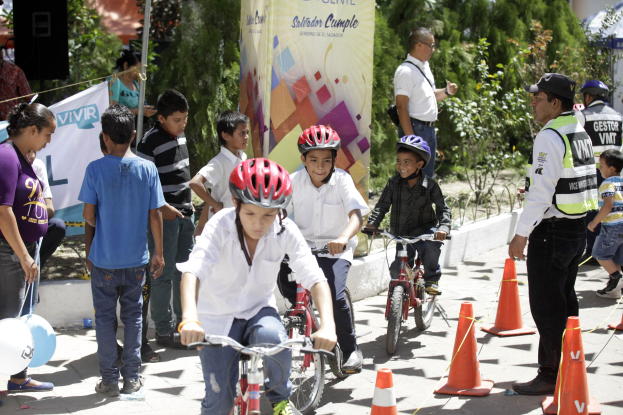 Kinder fahren mit Fahrrädern auf einer Straße mit Verkehrskegeln, einige tragen Helme, andere stehen daneben, mit einer Fahne, Bäumen und Gebäuden im Hintergrund.