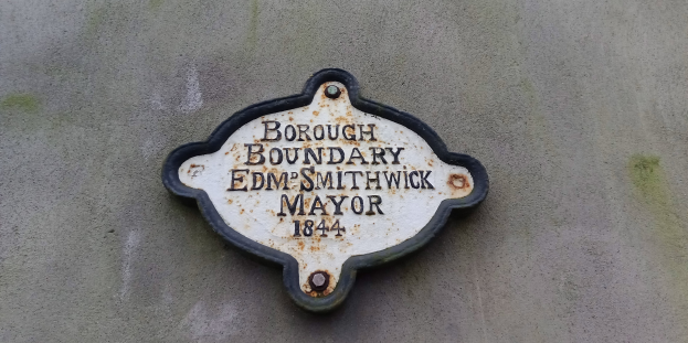 Metallplatte an der Außenseite eines Gebäudes mit schwarzem Hintergrund und weißer fettgedruckter Schrift, die "Gemarkungsgrenze Edm Smithwick Bürgermeister" liest.