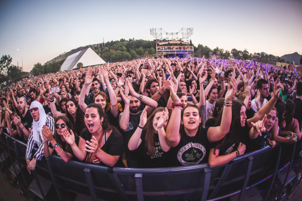 Menschenmenge auf einem Musikfestival mit erhobenen H├Ąnden vor einem Gel├Ąnder, mit B├Ąumen und Geb├Ąuden im Hintergrund bei klarem blauem Himmel.