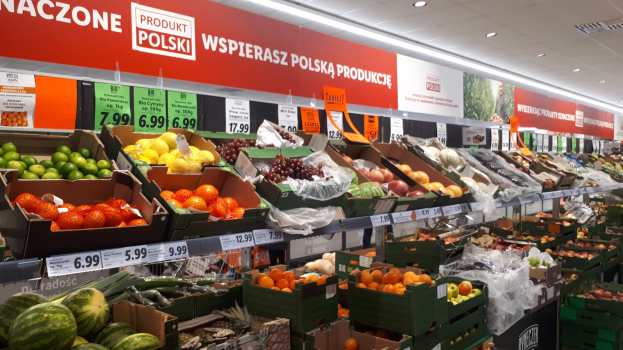 Ladeninneres mit verschiedenen Früchten und Gemüsen in Kartonschachteln mit Preisschildern, Wandtafeln mit Text und Bildern sowie Deckenbeleuchtung.