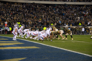 Ein Fußballspiel im Gange auf einem Stadionfeld mit Spielern in Sportkleidung und Helmen auf dem Boden, umgeben von Zuschauern und Stadionstrukturen.