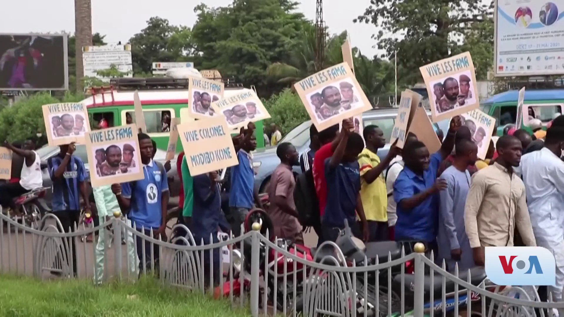 Eine Gruppe von Menschen mit Schildern steht vor einem Zaun, umgeben von Fahrzeugen, B├Ąumen, Masten, Werbetafeln und einem Bildschirm, mit Gras unten und Himmel im Hintergrund, scheinbar protestieren sie gegen eine Regierungentscheidung.