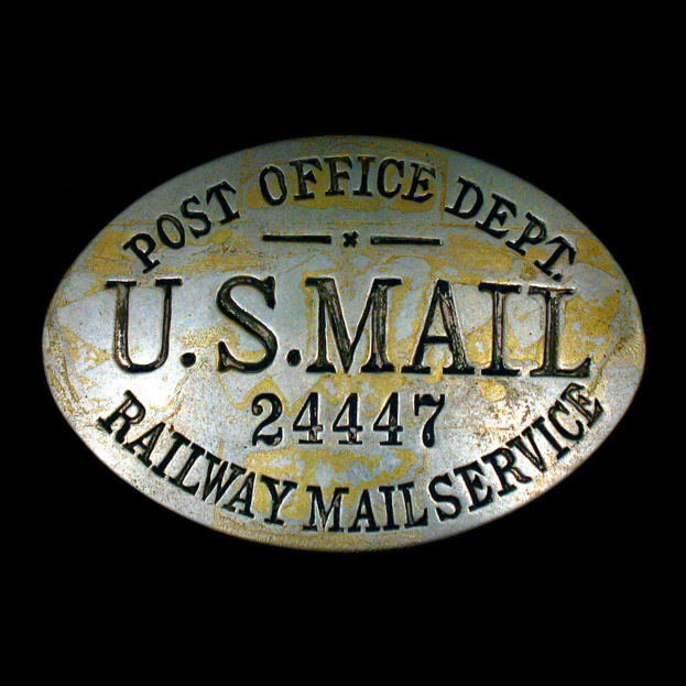 Ein ovalförmiges Metallobjekt mit der Aufschrift "Post Office Dept. U.S. Mail 2447 Railway Mail Service" auf einem schwarzen Hintergrund.