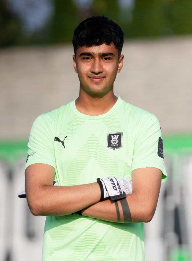 Ein junger Mann in einem grünen T-Shirt steht auf einem Fußballfeld und verschränkt die Arme, während er lächelt, wahrscheinlich ein Torwart, mit einem unscharfen Hintergrund.