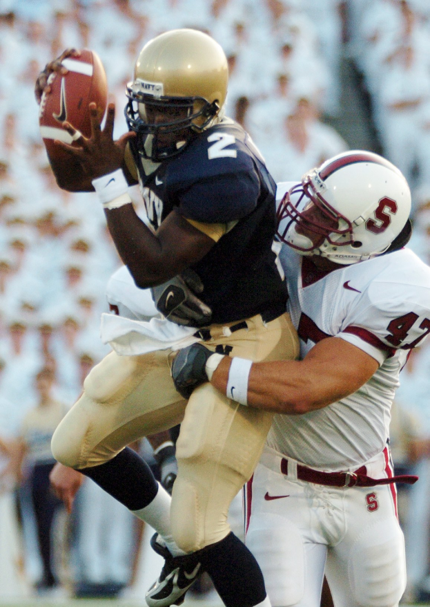 Zwei Fußballspieler in Helmen und Uniformen, einer fängt einen Ball in der Luft, während er von dem anderen getackelt wird, mit einem unscharfen Hintergrund.