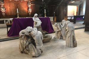 Eine Krippe in einer Kirche mit Statuen auf dem Boden, einem Tisch mit einem Tuch und Kerzenständern, Stühlen und einem geschmückten Weihnachtsbaum.