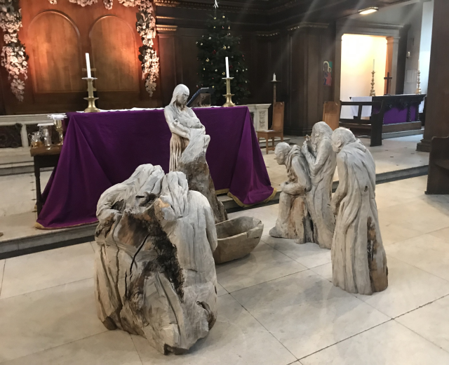 Eine Krippe in einer Kirche mit Statuen auf dem Boden, einem Tisch mit einem Tuch und Kerzenständern, Stühlen und einem geschmückten Weihnachtsbaum.