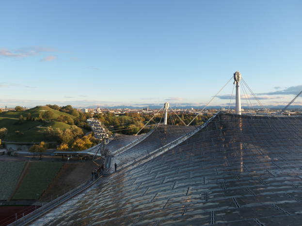 Das Olympiastadion in London, England, mit einem Dach voller Solarpanels, das eine Gruppe von Menschen auf dem Dach zeigt, einige Pfosten, einen Zaun, Gras, Pflanzen, Baumrinde, entfernte Gebäude und einen bewölkten Himmel.