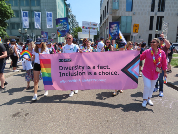 Eine Gruppe von Menschen marschiert auf einer Straße, hält ein Banner mit der Aufschrift "Diversität ist eine Tatsache, Inklusion ist eine Wahl", einige tragen Mützen und Schutzbrillen, andere halten zusätzliche Banner, vor einem Hintergrund aus Gebäuden, Bäumen, Laternenpfählen und einem klaren blauen Himmel.
