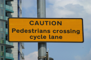 Gelbes Warnschild an einem Pfosten vor einem Gebäude, auf dem steht: "Fußgänger überqueren Fahrradspur" vor einem klaren blauen Himmel.