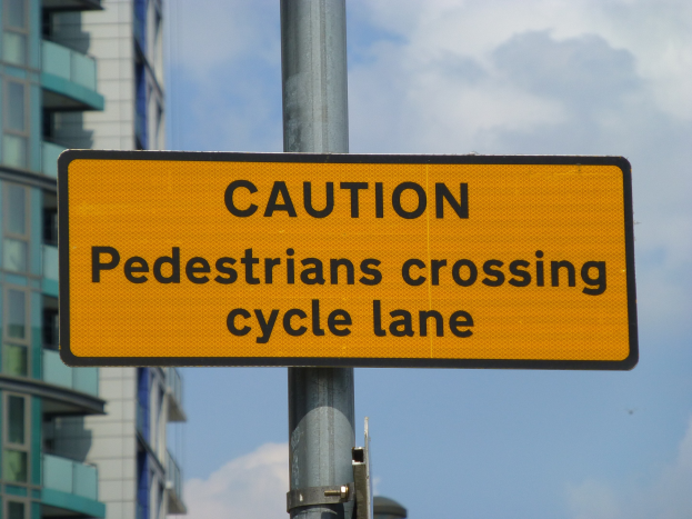 Gelbes Warnschild an einem Pfosten vor einem Gebäude, auf dem steht: "Fußgänger überqueren Fahrradspur" vor einem klaren blauen Himmel.