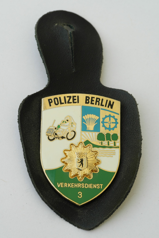 Ein schildförmiges Polizeibadge mit 'Polizei Berlin' in schwarzer fetter Schrift auf einem blauen Hintergrund mit einem weißen Stern und Rahmen, das auf einer Oberfläche ruht.
