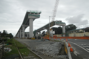 Baustelle mit einer Brücke im Hintergrund, eine Straße mit Absperrkegeln rechts, Steine und Gras am Boden, eine Bahnschiene links, Bäume und Gebäude auf beiden Seiten der Straße und ein bewölkter Himmel.