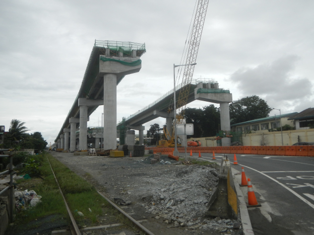 Baustelle mit einer Brücke im Hintergrund, eine Straße mit Absperrkegeln rechts, Steine und Gras am Boden, eine Bahnschiene links, Bäume und Gebäude auf beiden Seiten der Straße und ein bewölkter Himmel.