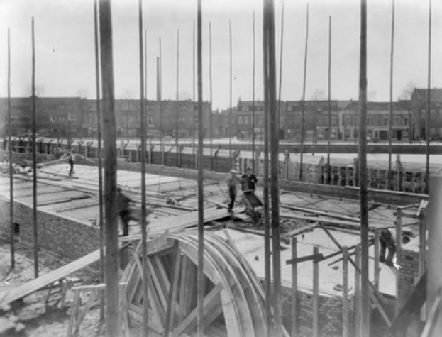 Schwarzes und weißes Foto einer Baustelle mit Arbeitern, hölzernen Pfählen und einem Rad im Vordergrund, Gebäuden, Bäumen und einem klaren Himmel im Hintergrund.