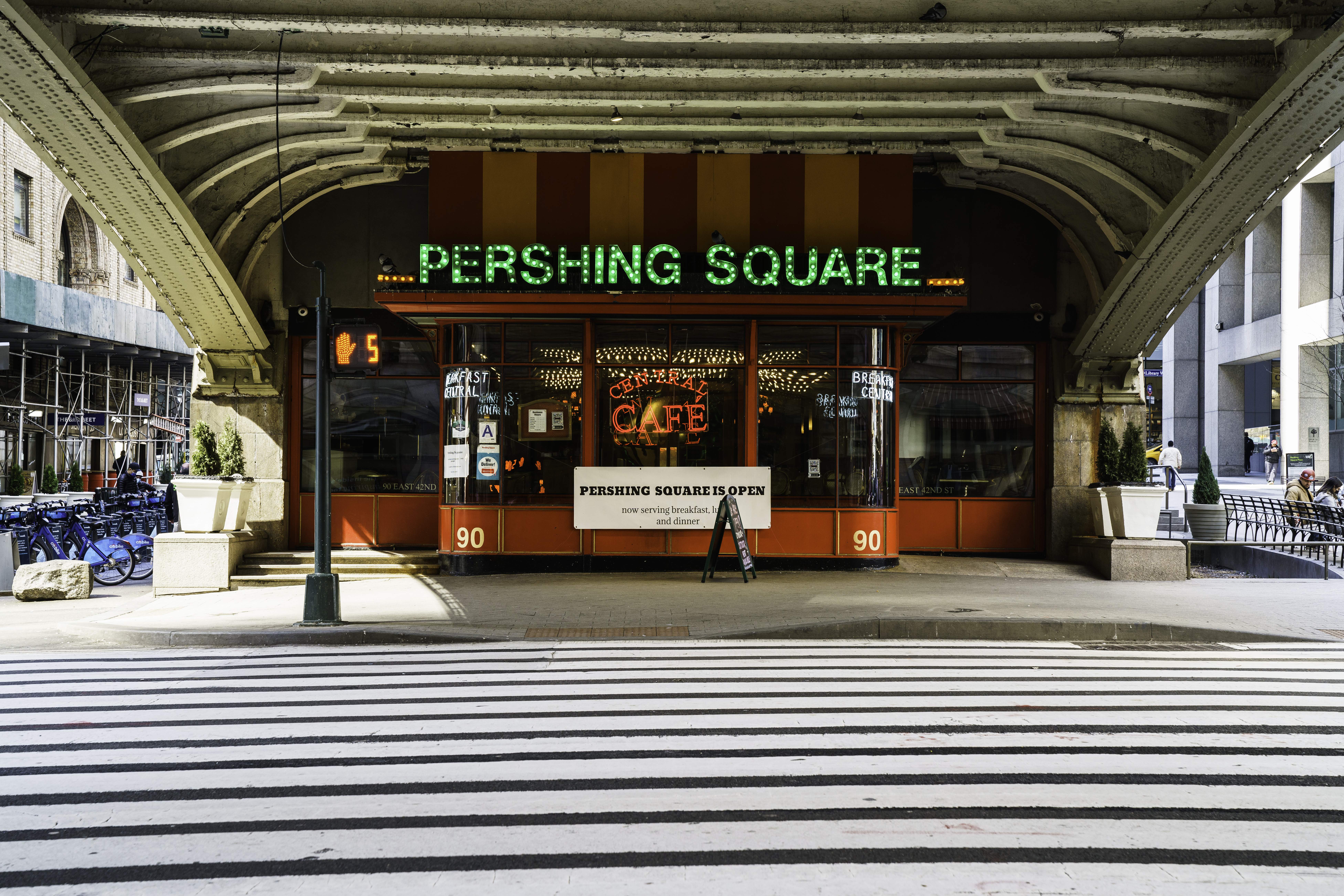 Eine belebte Stadtkulisse mit einem zentralen Gebäude, einem Schild mit der Aufschrift "Pershing Square Cafe in New York City", parkenden Fahrrädern, Topfpflanzen, einer Straßenlaterne, einem Metallzaun, Menschen auf Bänken sitzend, Bäumen und einem bewölkten Himmel.