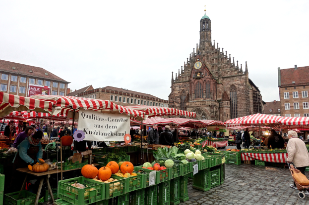 Ein belebter Markt in Nürnberg, Deutschland, mit verschiedenen Früchten und Gemüsen, Menschen, die umherlaufen, Zelten, Gebäuden mit Fenstern und einem Uhrenturm im Hintergrund bei einem sichtbaren Himmel.