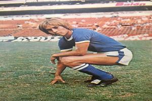 Ein Mann kniet auf einem Fußballfeld umgeben von Menschen auf Stühlen und Schildern mit Text, trägt Schuhe.