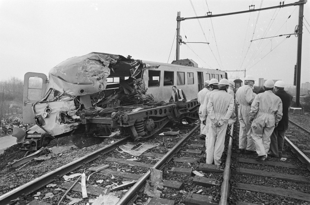 Eine Schwarz-Weiß-Szene einer Zugkollision mit einer Gruppe von Menschen in Helmen, die um das Wrack herumstehen, mit Strommasten, Bäumen, Gebäuden und einem klaren Himmel im Hintergrund.