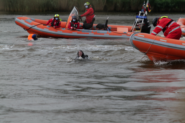 Gruppe von Menschen in einem Schlauchboot auf einem Fluss, mit zwei Personen im Wasser im Vordergrund und Vegetation im Hintergrund; alle tragen Schwimmwesten und Helme, was auf einen Rettungseinsatz hindeutet.