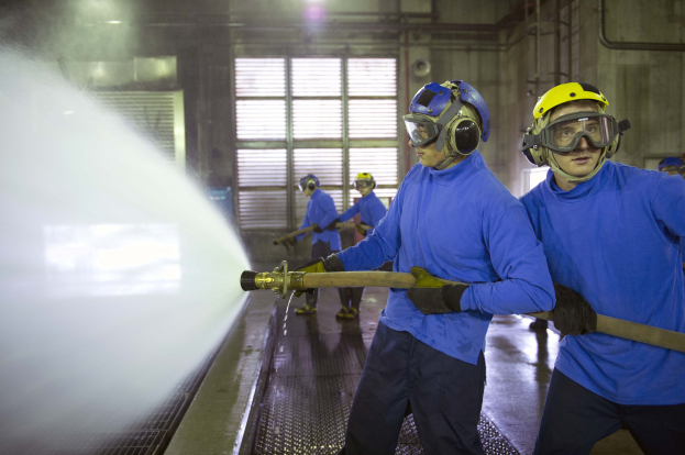 Gruppe von Männern in blauen Hemden und gelben Helmen, die an Maschinen arbeiten, wobei einer Wasser auf den Boden sprüht, in einer Fabrikumgebung.