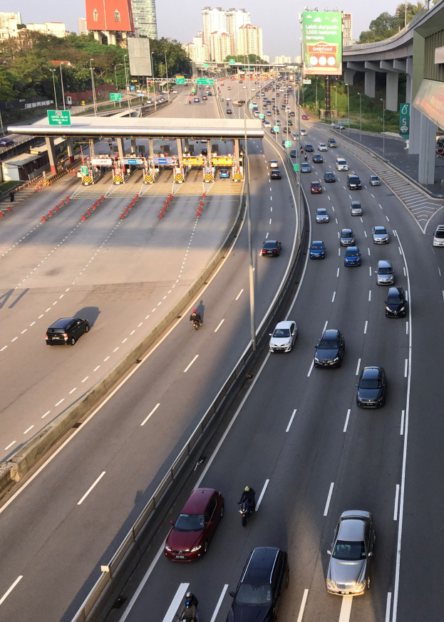 Eine belebte Autobahn mit Autos, Motorrädern, Verkehrskegeln, Verkehrszeichen, Laternen, einer Brücke, Bäumen, Gebäuden und einem klaren blauen Himmel.