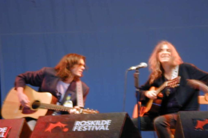 Zwei Männer auf der Bühne mit Musikinstrumenten, Mikrofon und 'ROCK SLIDE FESTIVAL' auf Kisten auf dem Boden.