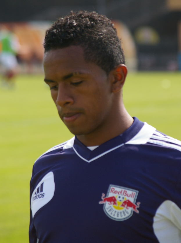 Ein junger Mann in einem blauen T-Shirt steht auf einem Fußballfeld, der als Flügelspieler mit kurzen dunklen Haaren und entschlossener Miene auf einem unscharfen Hintergrund zu sehen ist.