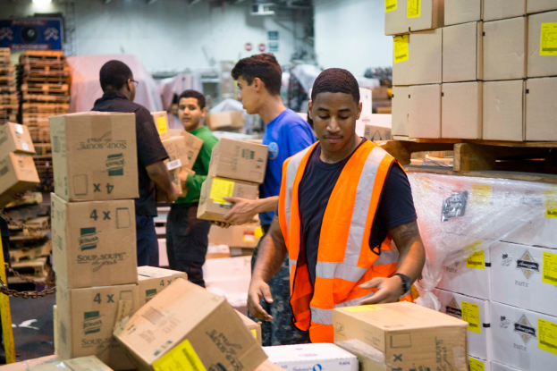 Eine Gruppe von Männern in orangen Westen und Sicherheitswesten lädt Kisten in ein Lager, mit mehreren Kartonagen auf der rechten Seite und einer Kette auf der linken Seite und einer Wand mit Lichtern oben im Hintergrund.