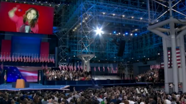 Eine große Menschenmenge sitzt vor einer Bühne mit einem Podium, einem Mikrofon, Flaggen, einem Bildschirm mit dem Bild einer Frau und Lichtausrüstung und wartet auf eine Präsidentschaftsdiskussion.