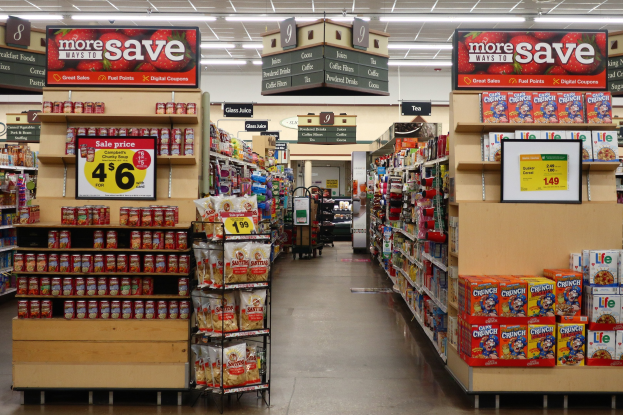 Ein Kiosk-Innenraum mit verschiedenen Lebensmitteln, die auf Gestellen, Regalen und dem Boden ausgebreitet sind, mit Deckenbeleuchtung und Informationsschildern und einer Person im Hintergrund.