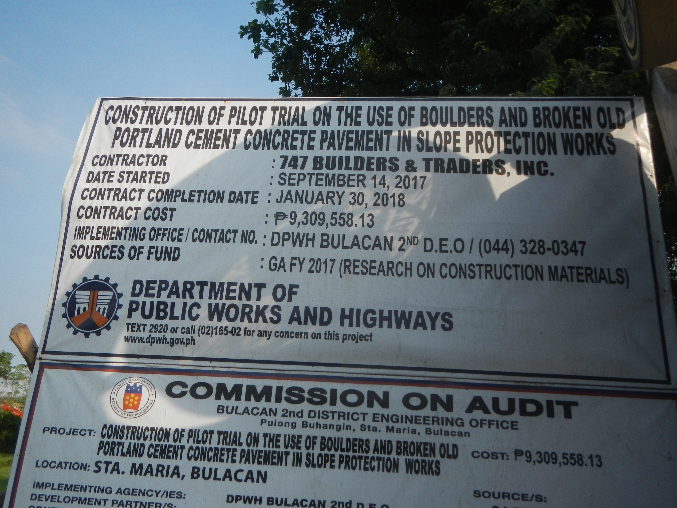 Schilder am Straßenrand mit der Aufschrift "Bauversuch zur Verwendung von Steinen und zerkleinertem Altbeton für den Schutz von Hängen" mit Bäumen, Pflanzen, Gras und einem klaren blauen Himmel im Hintergrund.